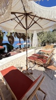A beach setting featuring woven umbrellas with fringe, casting shade over wooden lounge chairs with red cushions. A sandy surface leads towards the sea, which is visible in the background along with palm trees and additional seating. There is an overall tropical aesthetic with natural textures and a clear, sunny sky.