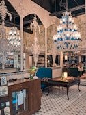 An ornately decorated interior with intricate white wall designs and multiple chandeliers featuring blue and clear glass elements. The space contains vintage furniture, including wooden cabinets and upholstered chairs. A patterned tile floor complements the detailed decor. There are potted plants and a glowing lamp on a table, adding warmth to the setting, while people are subtly visible in the background.