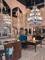 An ornately decorated interior with intricate white wall designs and multiple chandeliers featuring blue and clear glass elements. The space contains vintage furniture, including wooden cabinets and upholstered chairs. A patterned tile floor complements the detailed decor. There are potted plants and a glowing lamp on a table, adding warmth to the setting, while people are subtly visible in the background.