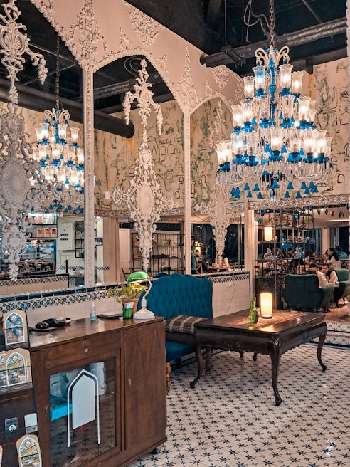 An ornately decorated interior with intricate white wall designs and multiple chandeliers featuring blue and clear glass elements. The space contains vintage furniture, including wooden cabinets and upholstered chairs. A patterned tile floor complements the detailed decor. There are potted plants and a glowing lamp on a table, adding warmth to the setting, while people are subtly visible in the background.