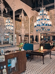 An ornately decorated interior with intricate white wall designs and multiple chandeliers featuring blue and clear glass elements. The space contains vintage furniture, including wooden cabinets and upholstered chairs. A patterned tile floor complements the detailed decor. There are potted plants and a glowing lamp on a table, adding warmth to the setting, while people are subtly visible in the background.