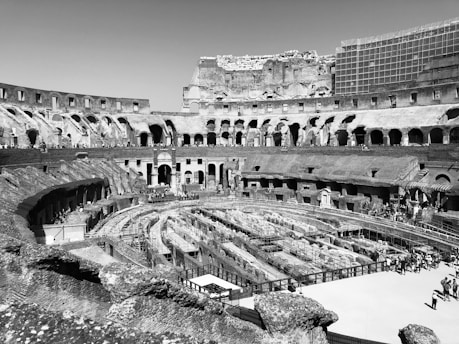 A historic amphitheater with multiple arches and a partially ruined structure. The interior shows a series of tiered seating areas with open spaces and columns. There are people walking and exploring the site, indicating it is a tourist destination. The overall structure is majestic, with a sense of ancient grandeur.