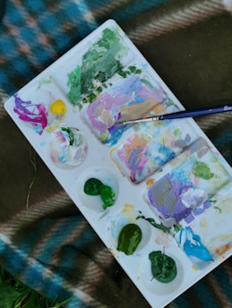 A paint palette with various colors including green, purple, pink, yellow, and blue is shown. The palette is placed on a checkered fabric with shades of blue and brown, and there is a paintbrush resting on the palette.