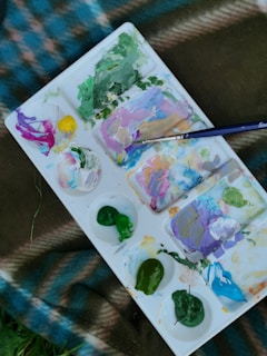 A paint palette with various colors including green, purple, pink, yellow, and blue is shown. The palette is placed on a checkered fabric with shades of blue and brown, and there is a paintbrush resting on the palette.