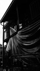 A black and white image of a building where a large sheet of plastic is draped over a balcony railing. The plastic catches the light, creating contrasting textures. The structure has metal poles and a wooden scaffold is visible below the balcony. The background includes a partly visible historical building with arched windows.
