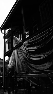 A black and white image of a building where a large sheet of plastic is draped over a balcony railing. The plastic catches the light, creating contrasting textures. The structure has metal poles and a wooden scaffold is visible below the balcony. The background includes a partly visible historical building with arched windows.