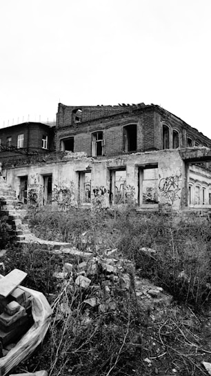 An abandoned and dilapidated brick building with broken windows and graffiti covering the walls. The surroundings are overgrown with wild vegetation and scattered debris, creating a sense of neglect.