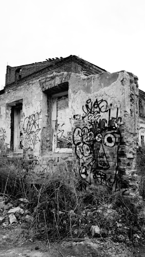 A dilapidated building with exposed brick and crumbling walls, covered in various graffiti. The structure looks abandoned, with wild plants growing around the base and rubble scattered on the ground. Black and white hues dominate the image, emphasizing the stark decay and urban art.