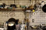 A well-organized kitchen with a variety of dishes stacked on a wall-mounted shelf. Glassware hangs upside down beneath the shelf, and several kitchen utensils are neatly arranged. A rice cooker, some containers, and cooking tools are on the countertop. The background features white subway tiles, adding to the clean and organized aesthetic.