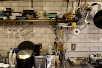 A well-organized kitchen with a variety of dishes stacked on a wall-mounted shelf. Glassware hangs upside down beneath the shelf, and several kitchen utensils are neatly arranged. A rice cooker, some containers, and cooking tools are on the countertop. The background features white subway tiles, adding to the clean and organized aesthetic.