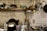 A well-organized kitchen with a variety of dishes stacked on a wall-mounted shelf. Glassware hangs upside down beneath the shelf, and several kitchen utensils are neatly arranged. A rice cooker, some containers, and cooking tools are on the countertop. The background features white subway tiles, adding to the clean and organized aesthetic.