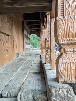 The image features a wooden walkway marked by intricately carved columns. The path leads into a verdant background, with the craftsmanship showcasing detailed patterns on the wood. The scene conveys an old-world charm with its rustic texture and natural setting.