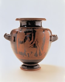 A large, ornate ancient Greek vase depicting detailed red-figure artwork on a black background. The central scene illustrates a group of people in various poses, possibly engaged in a mythological or ceremonial event. The vase has two handles, a broad body tapering to a narrow base, and intricate patterns around the rim.