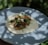 A fresh taco is placed on a light blue plate. The taco comprises a tortilla base topped with shredded meat, chopped onions, diced tomatoes, and fresh cilantro leaves. The scene is outdoors, with soft shadows of leaves, suggesting dappled sunlight.