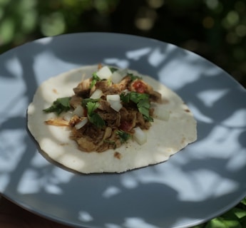 A fresh taco is placed on a light blue plate. The taco comprises a tortilla base topped with shredded meat, chopped onions, diced tomatoes, and fresh cilantro leaves. The scene is outdoors, with soft shadows of leaves, suggesting dappled sunlight.