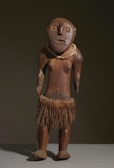 A wooden sculpture depicting a human-like figure with a simplistic and abstract design. It features a round head with large eyes, a flat nose, and a subtle mouth expression. The figure is adorned with a necklace and wears a skirt made of natural fibers, suggesting traditional or indigenous influence. The surface has a natural wood texture and a polished finish.