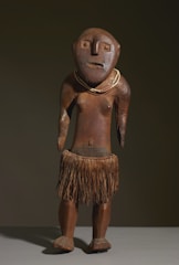 A wooden sculpture depicting a human-like figure with a simplistic and abstract design. It features a round head with large eyes, a flat nose, and a subtle mouth expression. The figure is adorned with a necklace and wears a skirt made of natural fibers, suggesting traditional or indigenous influence. The surface has a natural wood texture and a polished finish.