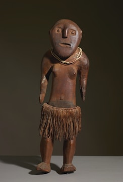 A wooden sculpture depicting a human-like figure with a simplistic and abstract design. It features a round head with large eyes, a flat nose, and a subtle mouth expression. The figure is adorned with a necklace and wears a skirt made of natural fibers, suggesting traditional or indigenous influence. The surface has a natural wood texture and a polished finish.