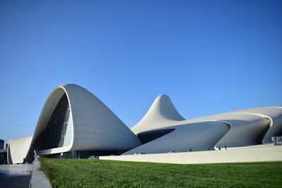 A modern architectural building with smooth, curving lines and an organic shape. The structure appears to be made of light-colored materials and features expansive glass windows. A well-manicured green lawn is visible in the foreground, while a clear blue sky stretches above. There are a few people walking near the building, giving a sense of scale to the large structure.