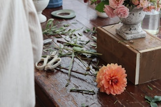 A rustic wooden table with various flower arranging tools and materials scattered around, including white pruning shears, green tape, and trimmed stems. A vibrant orange flower lies on the table next to a wooden block. In the background, there is an elegant vase with a floral arrangement featuring pink flowers.