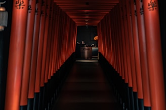 A dimly lit hallway lined with red columns, leading to a reception area with a desk that has 'KOMA' written on it. The design is reminiscent of traditional Japanese architecture, with a warm and inviting ambiance.