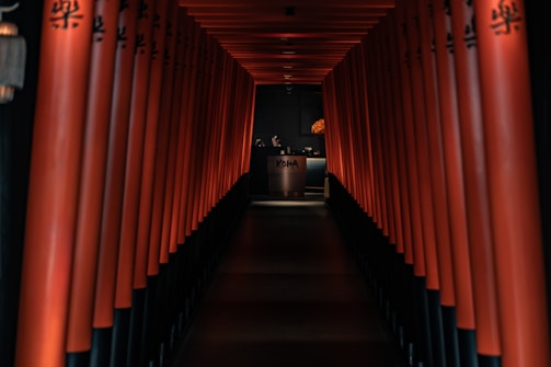 A dimly lit hallway lined with red columns, leading to a reception area with a desk that has 'KOMA' written on it. The design is reminiscent of traditional Japanese architecture, with a warm and inviting ambiance.