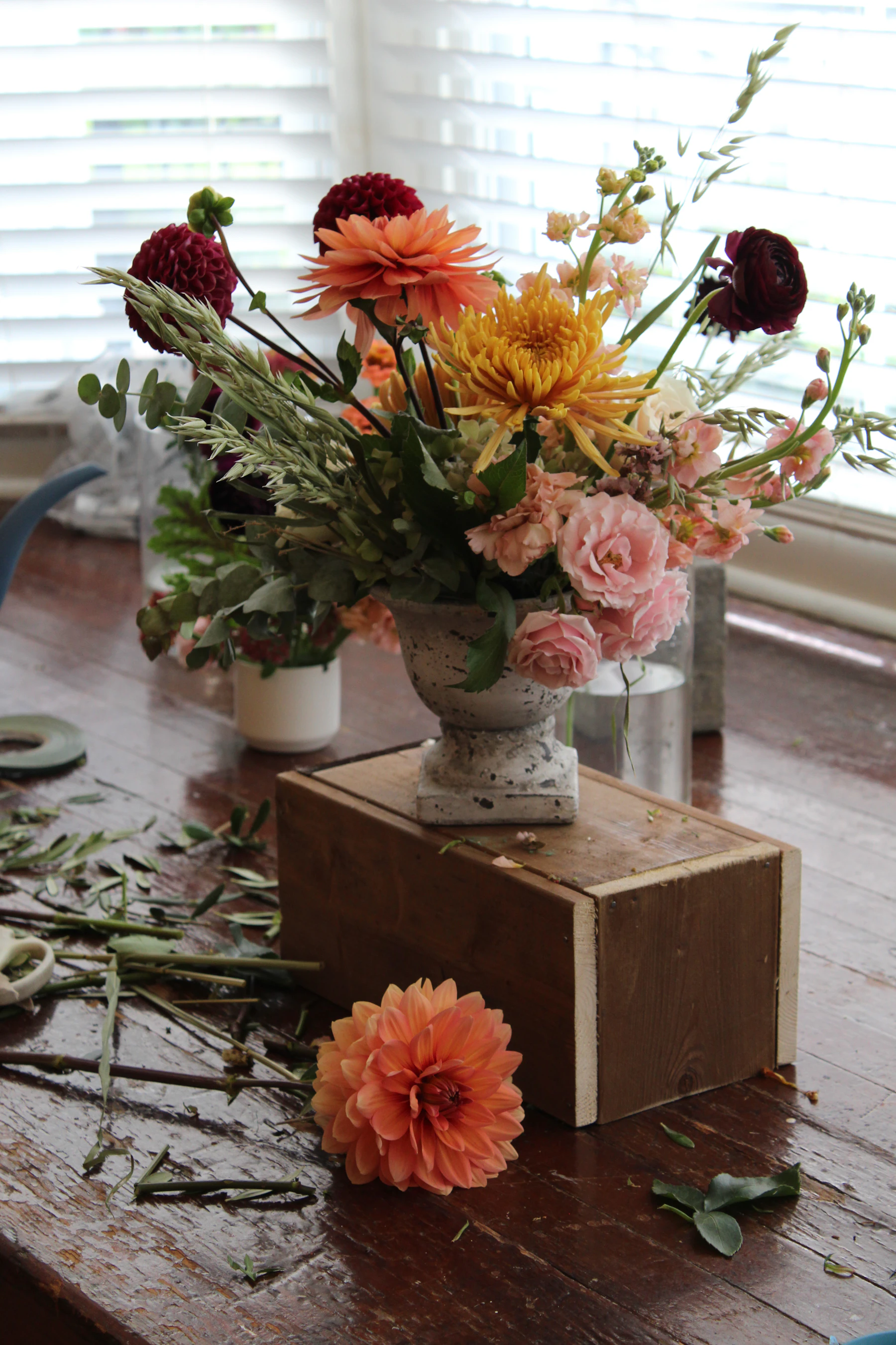 A footed urn of orange dahlias and pink roses on a wooden worktable scattered with stripped leaves and cut stem ends