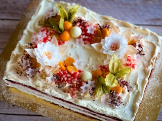 A beautifully decorated rectangular cake adorned with white frosting, topped with delicate white flowers, orange fruits, and light green leaves. Edible decorations like red and white sprinkles and small chocolate shavings enhance its elegance.