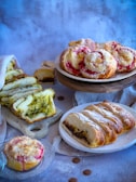 A variety of baked goods are displayed on different surfaces. There are slices of bread with green pesto filling placed on a wooden cutting board, round pastries with a pink filling and crumb topping on a wooden stand, and a braided loaf with a filling of chocolate or nuts on a white plate. The background is a muted blue and gray, adding a soft contrast to the warm tones of the baked items.