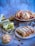 A variety of baked goods are displayed on different surfaces. There are slices of bread with green pesto filling placed on a wooden cutting board, round pastries with a pink filling and crumb topping on a wooden stand, and a braided loaf with a filling of chocolate or nuts on a white plate. The background is a muted blue and gray, adding a soft contrast to the warm tones of the baked items.