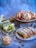 A variety of baked goods are displayed on different surfaces. There are slices of bread with green pesto filling placed on a wooden cutting board, round pastries with a pink filling and crumb topping on a wooden stand, and a braided loaf with a filling of chocolate or nuts on a white plate. The background is a muted blue and gray, adding a soft contrast to the warm tones of the baked items.