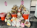 A large collection of trash and recycling items is piled up, consisting of cardboard boxes, plastic containers, and various packaging. The waste is overflowing from two large orange bins with pink signs above them, indicating different types of waste.