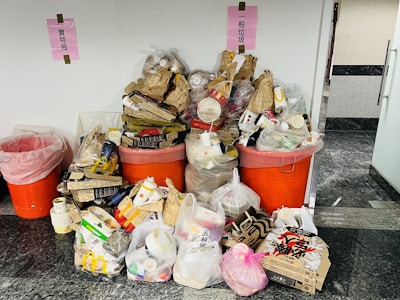 A large collection of trash and recycling items is piled up, consisting of cardboard boxes, plastic containers, and various packaging. The waste is overflowing from two large orange bins with pink signs above them, indicating different types of waste.