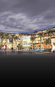 A modern shopping center with a glass facade, lit up under a cloudy sky. The complex is spacious with an open courtyard area featuring palm trees. People can be seen walking and sitting around, giving a sense of activity and liveliness. The architectural design appears sleek and contemporary, with multiple floors visible.