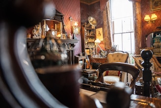 An intricately decorated room filled with ornate furniture, vintage decor, and antiques. The setting includes a fireplace with a mantel displaying various trinkets, a collection of bottles and jars, and framed artworks. Velvet drapes adorn a large window, allowing natural light to filter into the room, creating a warm ambiance.