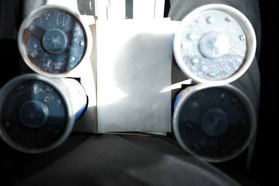 A cardboard drink holder carrying four plastic cups with lids, featuring condensation drops. The drinks are placed on a person's lap, wearing gray pants. Sunlight creates a dramatic contrast with shadows in the image.