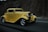 A vintage yellow hot rod car is driving on a highway, surrounded by a rocky landscape. The car features chrome wheels, a sleek body design, and prominent headlights, giving it an iconic, classic appearance.