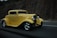 A vintage yellow hot rod car is driving on a highway, surrounded by a rocky landscape. The car features chrome wheels, a sleek body design, and prominent headlights, giving it an iconic, classic appearance.