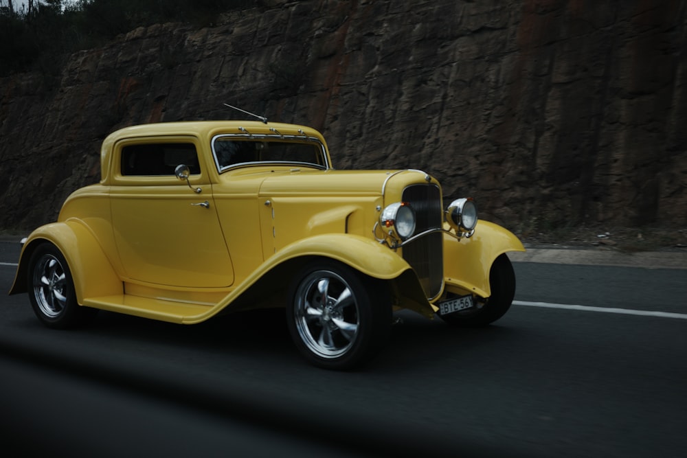 an old yellow car driving down the road