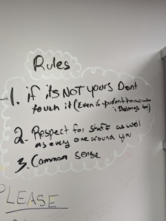 A whiteboard with three handwritten rules. The rules emphasize not touching things that aren't yours, showing respect for staff and others, and using common sense. Drawn with a black marker, the text is surrounded by simple, cloud-like sketches. Additional faint writing is visible around the main text.