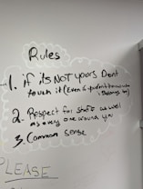 A whiteboard with three handwritten rules. The rules emphasize not touching things that aren't yours, showing respect for staff and others, and using common sense. Drawn with a black marker, the text is surrounded by simple, cloud-like sketches. Additional faint writing is visible around the main text.