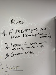 A whiteboard with three handwritten rules. The rules emphasize not touching things that aren't yours, showing respect for staff and others, and using common sense. Drawn with a black marker, the text is surrounded by simple, cloud-like sketches. Additional faint writing is visible around the main text.