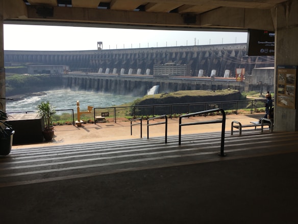 A large hydroelectric dam stands in the background with water flowing into a reservoir. The scene is viewed from a covered observation area with steps and railings leading down to a walkway. There are plants in pots and informational signs present.