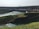 A massive hydroelectric dam spans across a wide river with water flowing from its openings. The foreground features lush green grass and vegetation covering the banks, while a flow of water is seen moving towards the dam structure.