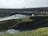 A massive hydroelectric dam spans across a wide river with water flowing from its openings. The foreground features lush green grass and vegetation covering the banks, while a flow of water is seen moving towards the dam structure.
