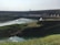 A massive hydroelectric dam spans across a wide river with water flowing from its openings. The foreground features lush green grass and vegetation covering the banks, while a flow of water is seen moving towards the dam structure.