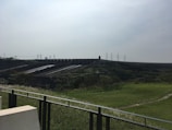 A large dam structure stretches across the landscape, with multiple sluice gates and channels. The foreground includes grassy areas and a metal fence, while electricity pylons are visible in the background against a cloudy sky.