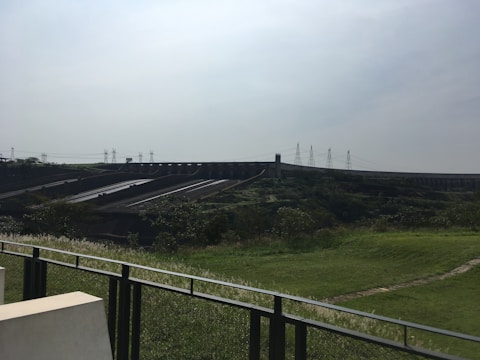 A large dam structure stretches across the landscape, with multiple sluice gates and channels. The foreground includes grassy areas and a metal fence, while electricity pylons are visible in the background against a cloudy sky.