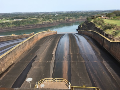 A large dam structure with sloped concrete surfaces leading to a body of water. Vegetation and green fields stretch across the horizon with distant trees and buildings. Guard rails and metal railings are visible near the bottom of the dam.