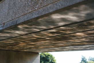 The image depicts a concrete structure, possibly an underside of a bridge, with visible light reflections creating a pattern on the surface. The background has some blurred greenery, suggesting an outdoor setting.